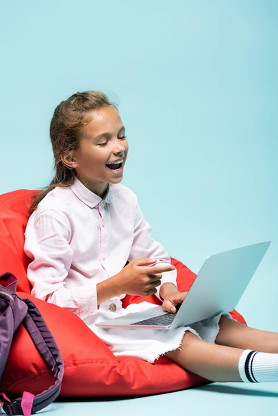 Laughing schoolgirl pointing at laptop on blue background  - Foto, imagen