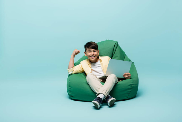 Smiling asian schoolkid showing yes gesture and holding laptop on beanbag chair on blue background  - Fotó, kép