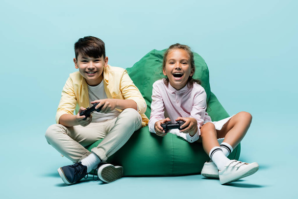 KYIV, UKRAINE - JULY 2, 2021: Happy multiethnic preteen kids playing video game on beanbag chair on blue background  - Φωτογραφία, εικόνα