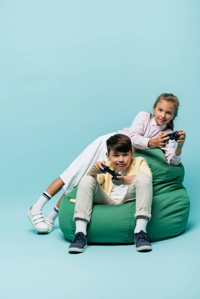 KYIV, UKRAINE - JULY 2, 2021: Multiethnic children playing video game on beanbag chair on blue background  - Fotografie, Obrázek