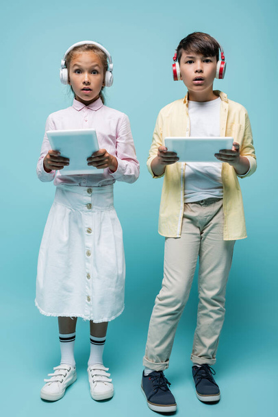 Écoliers interracial choqués dans des écouteurs tenant des tablettes numériques sur bleu  - Photo, image