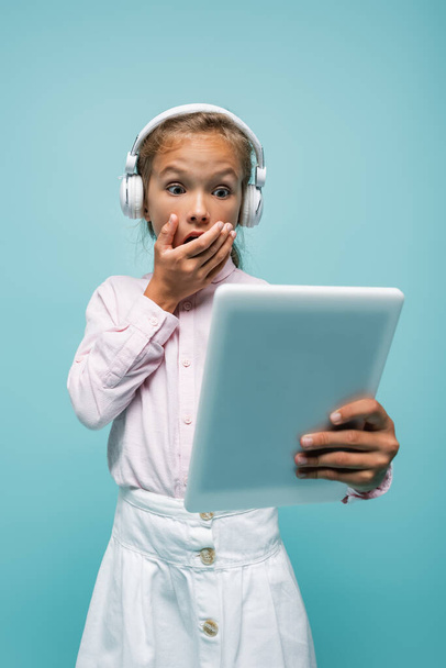 Shocked schoolgirl in headphones looking at digital tablet isolated on blue  - Photo, Image