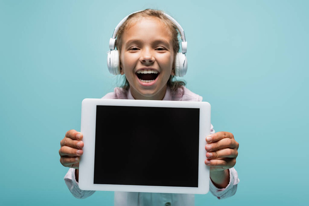 Smiling schoolgirl in headphones looking at camera and holding digital tablet isolated on blue  - Fotó, kép