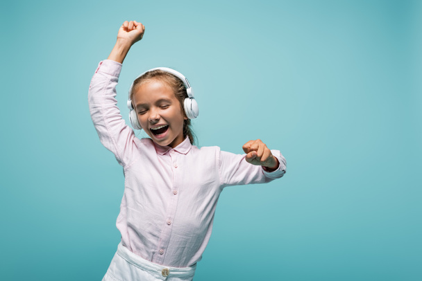 Cheerful schoolkid listening music in headphones isolated on blue  - 写真・画像