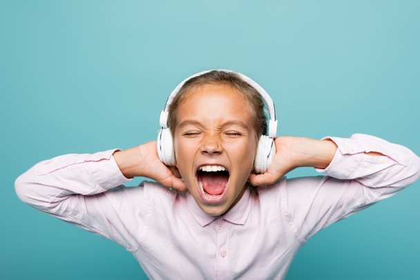 Angry preteen schoolgirl listening music in headphones isolated on blue  - Foto, imagen