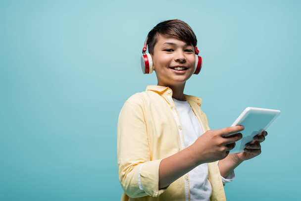 Smiling asian schoolkid in headphones holding digital tablet isolated on blue  - Фото, изображение
