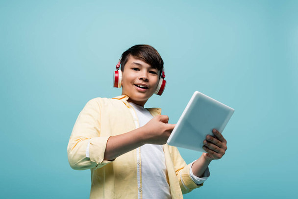 Low angle view of smiling asian schoolboy in headphones holding digital tablet isolated on blue  - Fotografie, Obrázek