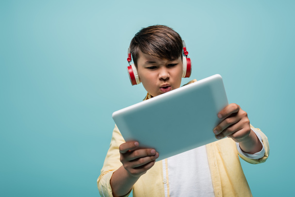 Focused asian schoolkid in headphones using digital tablet isolated on blue  - Photo, Image