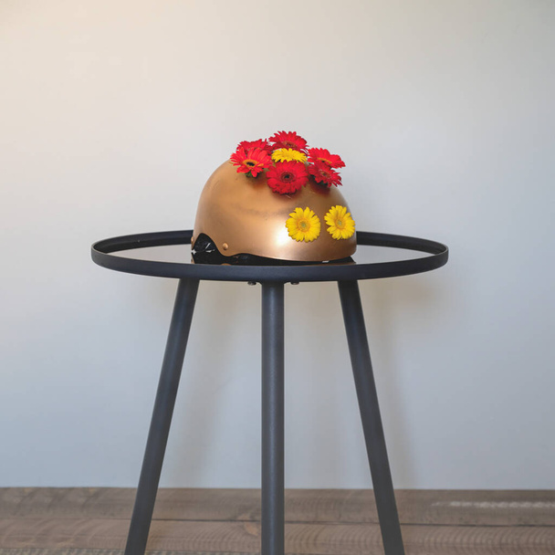 Домашняя ваза из шлема цвета золота. Вторая жизнь неиспользованных вещей. Цветы Герберы для домашнего декора. Золотая ваза на черном маленьком круглом столе - Фото, изображение