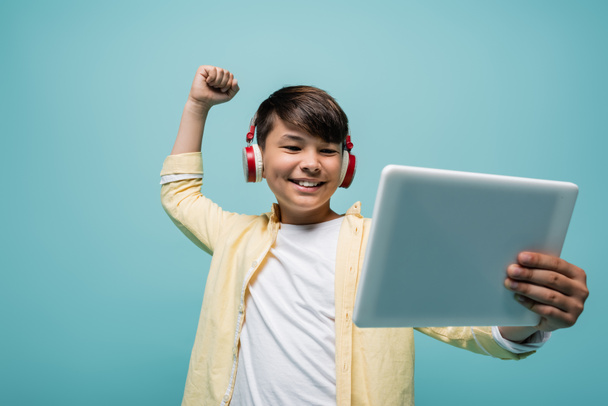 Positive asian schoolboy in headphones showing yes gesture and holding digital tablet isolated on blue  - Photo, Image