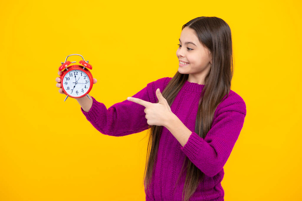 Возбужденное лицо. Ребенок держит часы изолированными на желтом студийном фоне. Подросток с будильником, показывающим время. Удивительное выражение лица, жизнерадостное и радостное - Фото, изображение