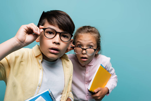 Focused interracial schoolkids in eyeglasses holding notebooks and looking at camera isolated on blue  - Фото, зображення