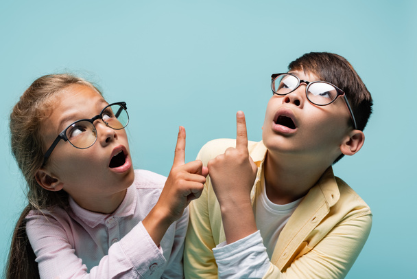 Shocked interracial schoolkids in eyeglasses pointing with fingers isolated on blue  - Фото, зображення