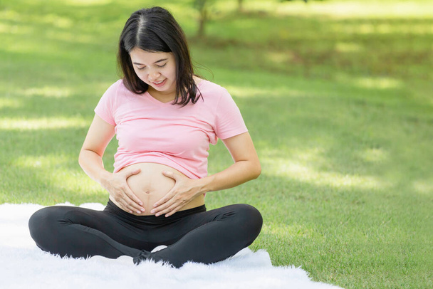 Ευτυχισμένη έγκυος γυναίκα κάθεται να χαλαρώσετε στο πάρκο και να κάνει χειρονομία καρδιάς - Φωτογραφία, εικόνα