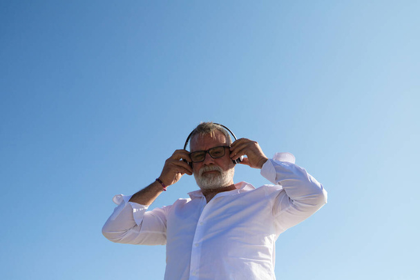 handsome mature man with beard and grey hair is in the park while listening to music with his headphones. Picture taken from below and the blue sky in the background. Senior concept, travel and music. - 写真・画像