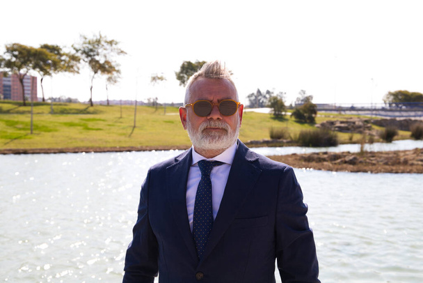 Homme mûr beau, exécutif, cheveux gris, barbe, lunettes de soleil, veste et cravate, est dans un parc avec un lac artificiel posant pour la photo. Concept d'homme mûr, grand-père, exécutif. - Photo, image