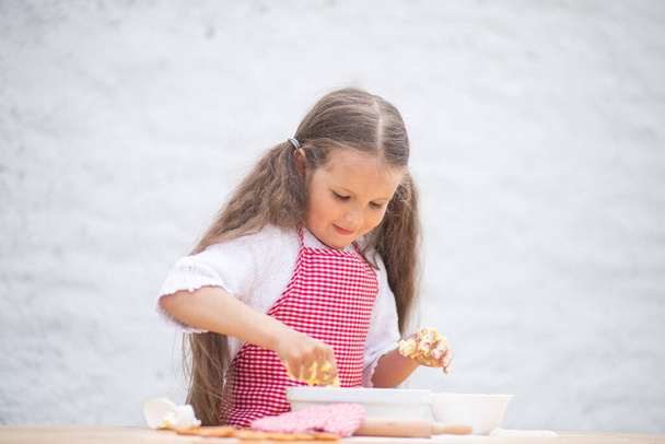 シェフのコスチュームで幸せな少女は,生地をひざまずいてパイを調理するのに役立ちます, 子供の笑顔, レシピに応じて調理, 女の子は,ママのために料理するのに役立ちます, 子供の肖像画と開発. - 写真・画像