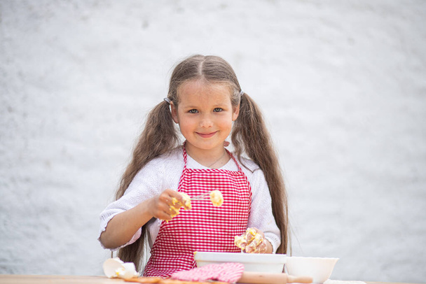 Šťastná holčička v kostýmu kuchaře pomáhá uvařit koláč hnětením těsta, dětský úsměv, vaření podle receptu, dívka pomáhá vařit pro mámu, dětský portrét a vývoj. - Fotografie, Obrázek