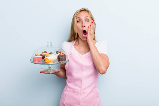 blonde young adult woman feeling shocked and scared. home made cakes concept - Foto, Imagen