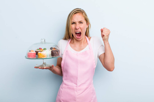 blonde young adult woman shouting aggressively with an angry expression. home made cakes concept - Foto, Imagen
