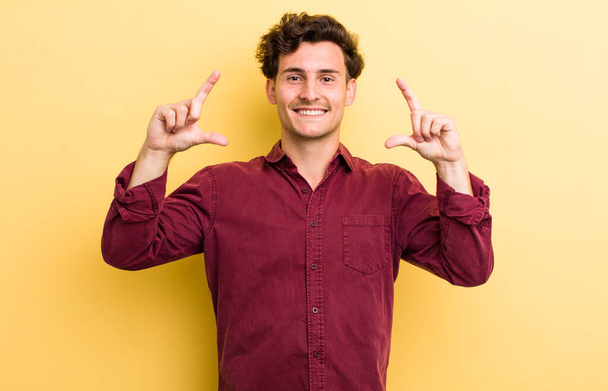young handsome man framing or outlining own smile with both hands, looking positive and happy, wellness concept - 写真・画像
