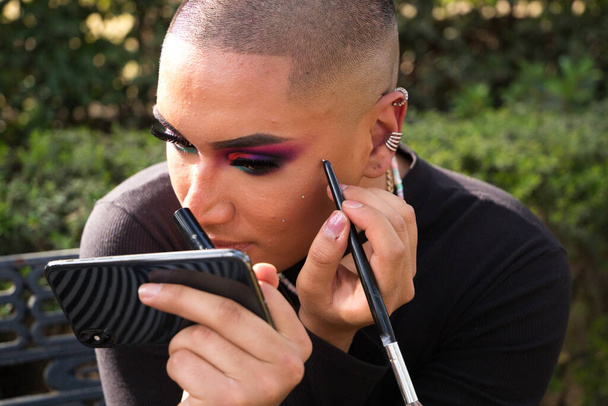 Jonge niet-binaire persoon uit Zuid-Amerika doet make-up op terwijl ze naar haar mobiele telefoon kijkt. de persoon draagt make-up en zwarte kleren. Begrip homoseksualiteit, mode en schoonheid. - Foto, afbeelding
