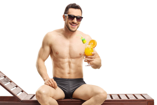 Young man sitting on a sunbed and holding a cocktail isolated on white background - Foto, Imagen