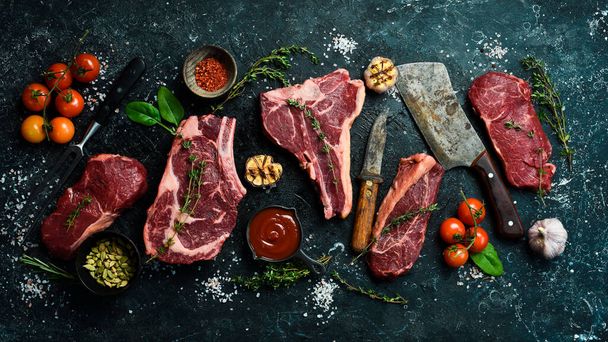 Variety of raw aged beef steaks: t-bone, tomahawk, striploin, tenderloin, new york steak for grilling with spices on stone background. On a black stone background. - 写真・画像