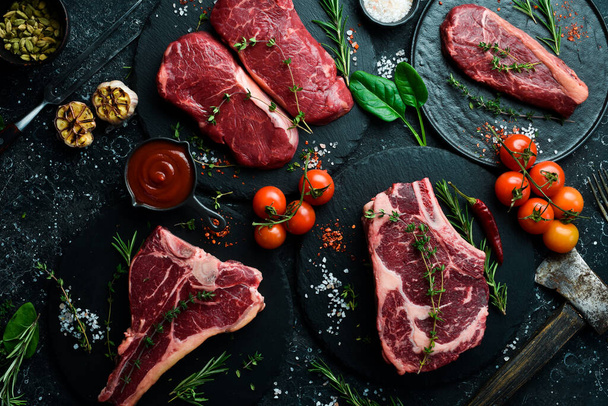A variety of raw beef steaks for grilling with seasonings: T-bone, striploin steak, ribeye. Top view. On a black stone background. - 写真・画像