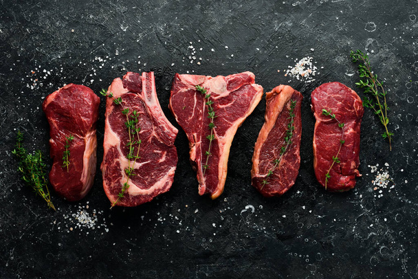 A variety of raw beef steaks for grilling with seasonings: T-bone, striploin steak, ribeye. Top view. On a black stone background. - Zdjęcie, obraz