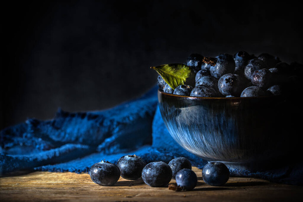 Blueberries in a blue bowl with napkin on wooden board table and background - dark and moody - Foto, Bild