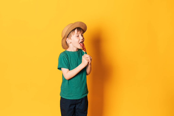 Boy in hat licks lollipop in the shape of piece of watermelon against yellow background. - Photo, Image