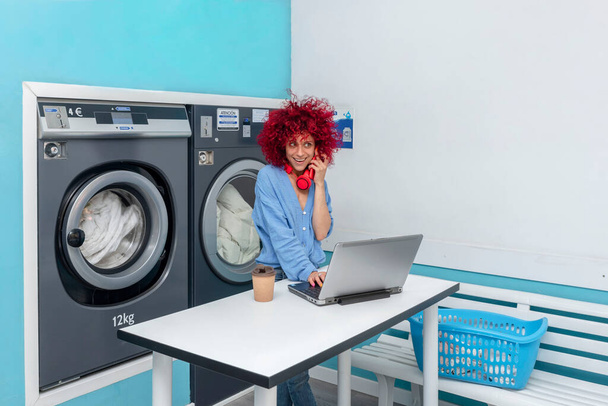 usmívající se mladá latina žena s červenými afro vlasy pracuje se svým notebookem a mluví do telefonu v modré prádelně, zatímco čeká na prádlo, její červené sluchátka kolem krku, šálek kávy na stole - Fotografie, Obrázek