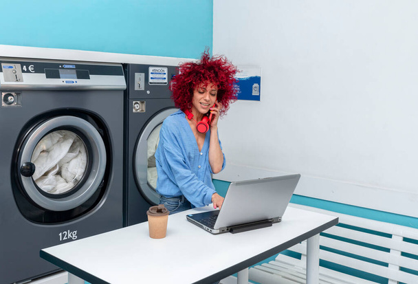 une jeune femme latina souriante aux cheveux roux afro travaille avec son ordinateur portable et parle au téléphone dans la buanderie bleue, en attendant que la lessive soit faite, ses écouteurs rouges autour du cou, une tasse de café sur la table - Photo, image