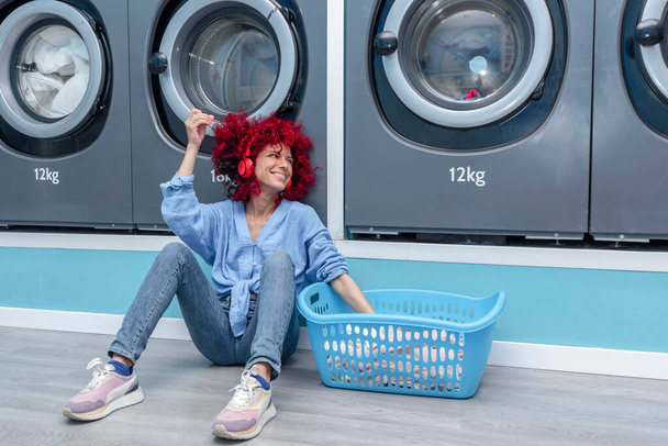 Una joven latina sonriente con el pelo rojo afro sentada en una lavandería automática azul escuchando música con auriculares rojos mientras espera a que se haga la colada. - Foto, Imagen