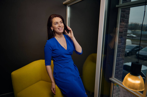 Πορτρέτο μιας μοντέρνας καυκάσιας γυναίκας που κάθεται στην κίτρινη πολυθρόνα και μιλάει στο κινητό της τηλέφωνο σε ένα γραφείο εσωτερικού σχεδιασμού. Κομψή επιχειρηματίας, διακοσμητής, κτηματομεσίτης - Φωτογραφία, εικόνα