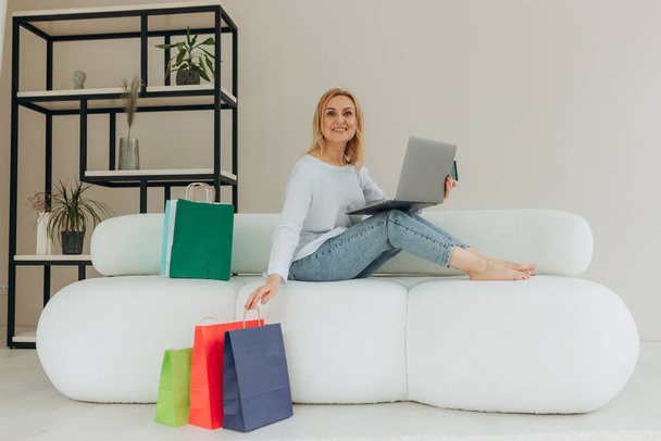 Joyful pretty caucasian mature blond stylish woman, happy with new purchased clothes, sitting on a sofa at home, using laptop and credit card for online payments, many paper bags near, smiling - Foto, imagen
