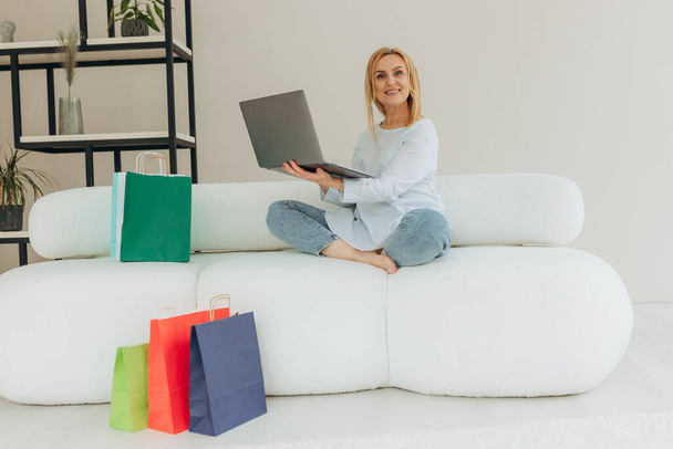 Joyful pretty caucasian mature blond stylish woman, happy with new purchased clothes, sitting on a sofa at home, using laptop and credit card for online payments, many paper bags near, smiling - Фото, зображення