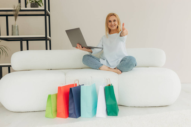 Portré érett vonzó csinos csinos, vidám nő ül a kanapén segítségével laptop vásárlás. A nő boldog vásárlás mellett a sok bevásárló táska otthon. Nő mutatja osztály hüvelykujját - Fotó, kép