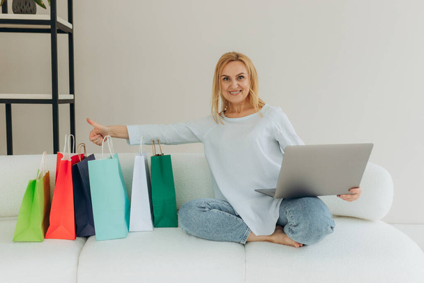 Mujer madura de compras en línea en casa en el ordenador portátil. La mujer hace un pedido en línea. Mujer atractiva con bolsas de compras. Concepto de compras online - Foto, Imagen