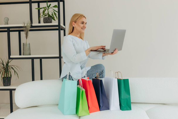 Mujer madura de compras en línea en casa en el ordenador portátil. La mujer hace un pedido en línea. Mujer atractiva con bolsas de compras. Concepto de compras online - Foto, imagen