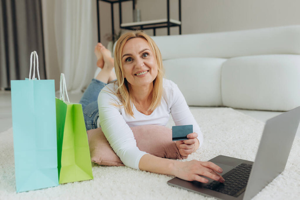 Happy mature woman holding credit card, using laptop pc, choosing and buying goods on web, lying on floor indoors - Photo, image