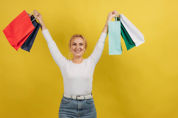 Šťastná dospělá žena zvedá ruce s nákupy na izolovaném žlutém pozadí. Žena drží barevné tašky. - Fotografie, Obrázek