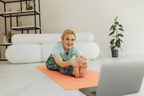 Senior woman sitting on the floor on exercise mat in front of laptop and training at home. Beautiful senior woman doing stretching exercise while sitting on yoga mat at home. - Photo, Image