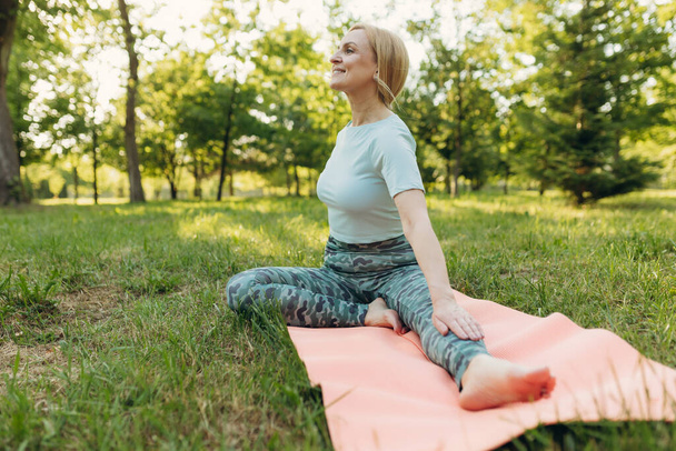 Orta yaşlı bir kadın yoga egzersizi sırasında yeşil çimlerde dinleniyor, sabah rutini, gün batımı ışığı, açık havada. Mavi spor kıyafetli olgun bir kadın esneme hareketleri yapıyor. Yaş, sağlıklı yaşam tarzı. - Fotoğraf, Görsel