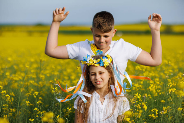 Caring kind big older brother braids multi-colored thin long ribbons in bright Ukrainian flower wreath on head of little smiling younger sister, in blooming rapeseed endless field under clear blue sky - Fotó, kép