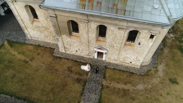 Eine junge Braut im Brautkleid nähert sich dem Bräutigam und umarmt ihn. Vor dem Hintergrund der Mauern der alten Kirche. - Filmmaterial, Video
