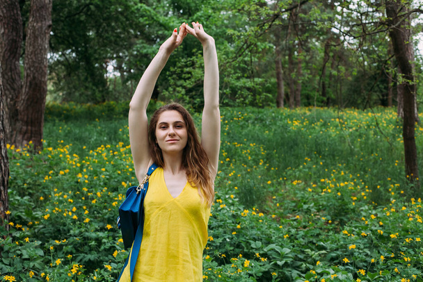 Молодая девушка с кудрявыми светлыми волосами в жёлтом летнем платье и голубой сумочке стоит на цветущей лужайке в лесу, поднимает руки и смеется.. - Фото, изображение