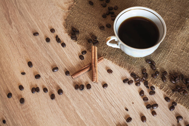 Ένα φλιτζάνι ευωδιαστό μαύρο καφέ, λινό ύφασμα και ξυλάκια κανέλας βρίσκονται σε ένα ξύλινο καφέ τραπέζι. Οι κόκκοι του καφέ είναι διάσπαρτοι στην επιφάνεια του τραπεζιού.. - Φωτογραφία, εικόνα