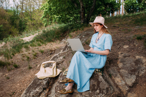 Eine junge Freiberuflerin arbeitet in der Natur mit einem Laptop. Bekleidet mit einem blauen Sommerkleid und einem hellen Hut. Sitzt auf Steinen in einem Waldgebiet. In der Nähe liegt eine beige Ledertasche. - Foto, Bild